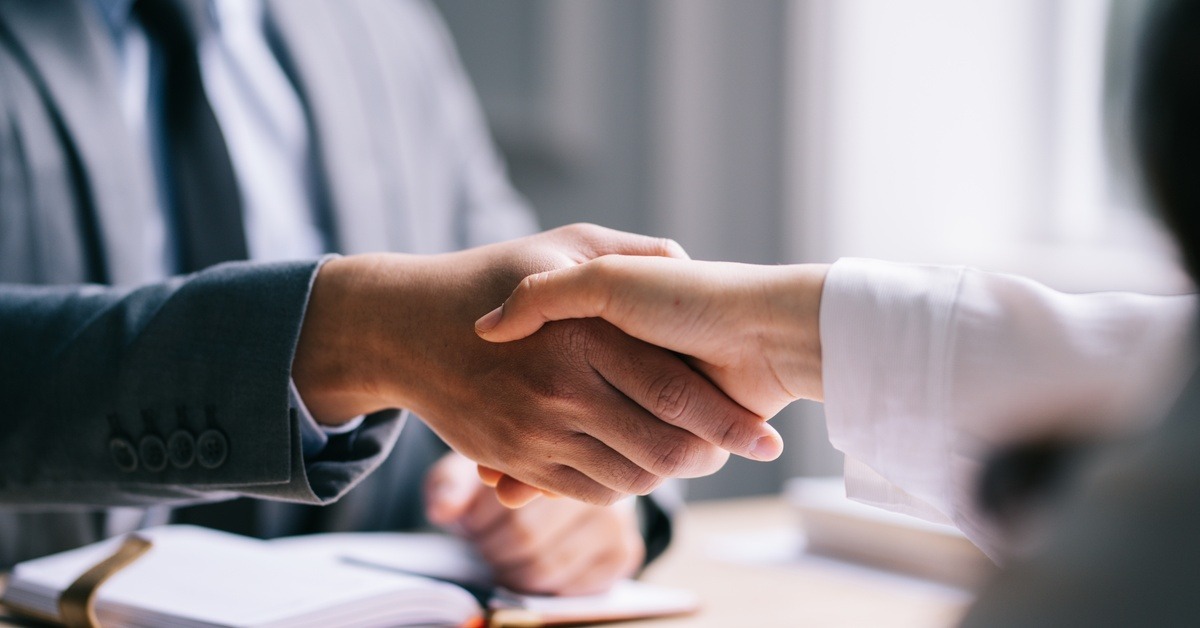 A close-up of two people in business-professional attire shaking hands over a desk and some paperwork.