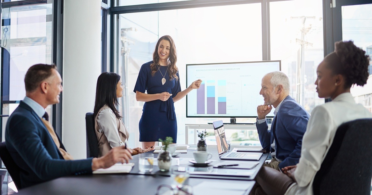 A boardroom meeting of five people being led by a woman pointing to a TV screen that displays chart data.