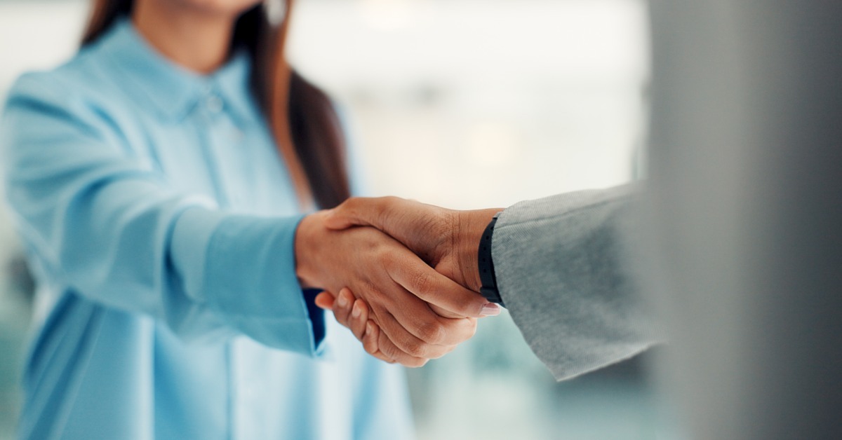 A woman in a light blue blouse shakes hands with someone in a gray suit, with a blurred office behind.