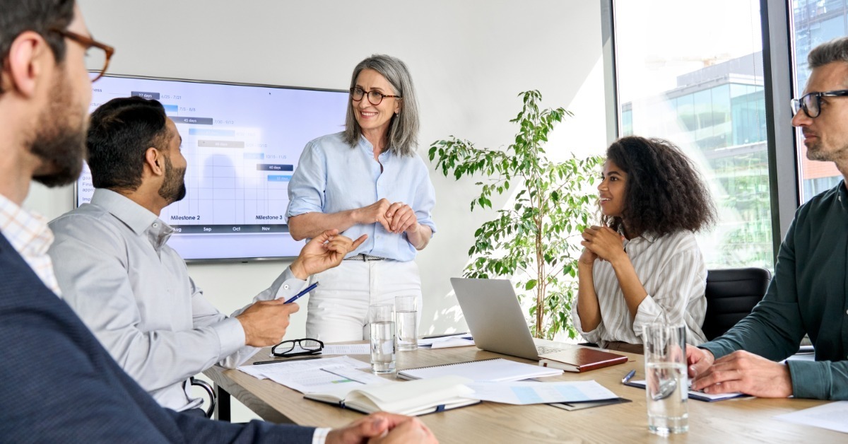 A woman stands by a screen while four coworkers sit at a conference table with laptops, notes, and water glasses.