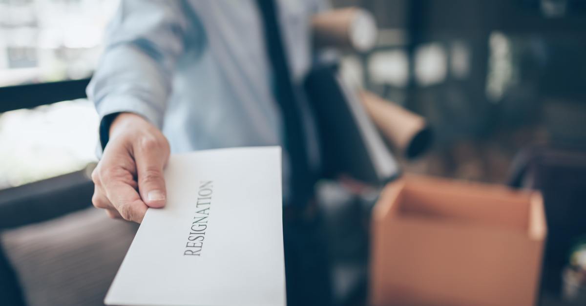 A man in a suit hands over a resignation letter with one arm while carrying office items under the other arm.