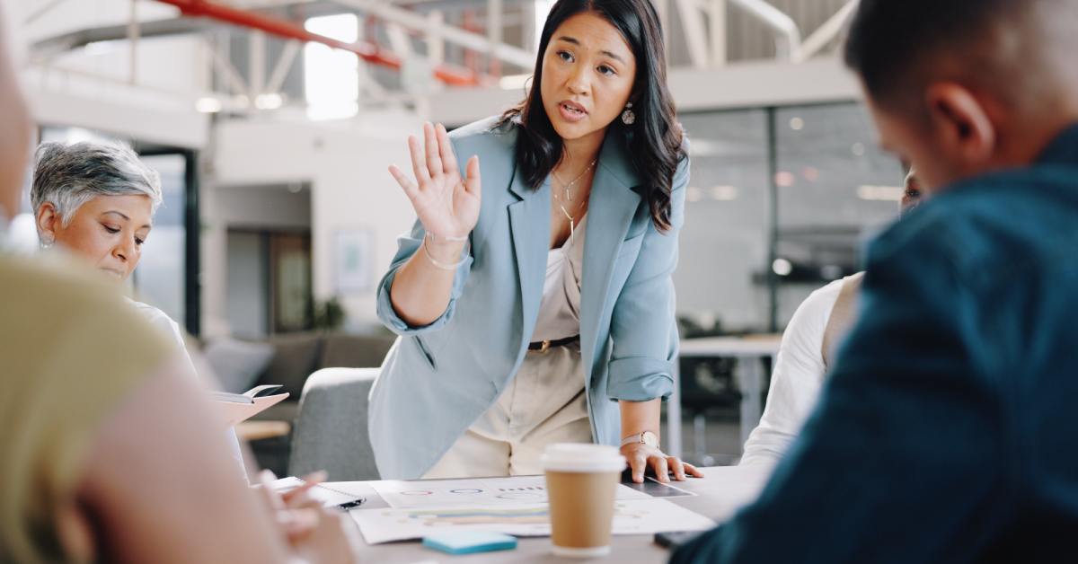 A woman in a light blue blazer holds her hand up while leading a team discussion around a table in a modern office.