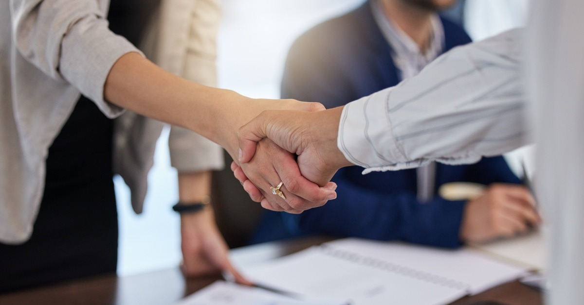 A close-up of two people shaking hands over paperwork on a desk, with another person blurred in the background.