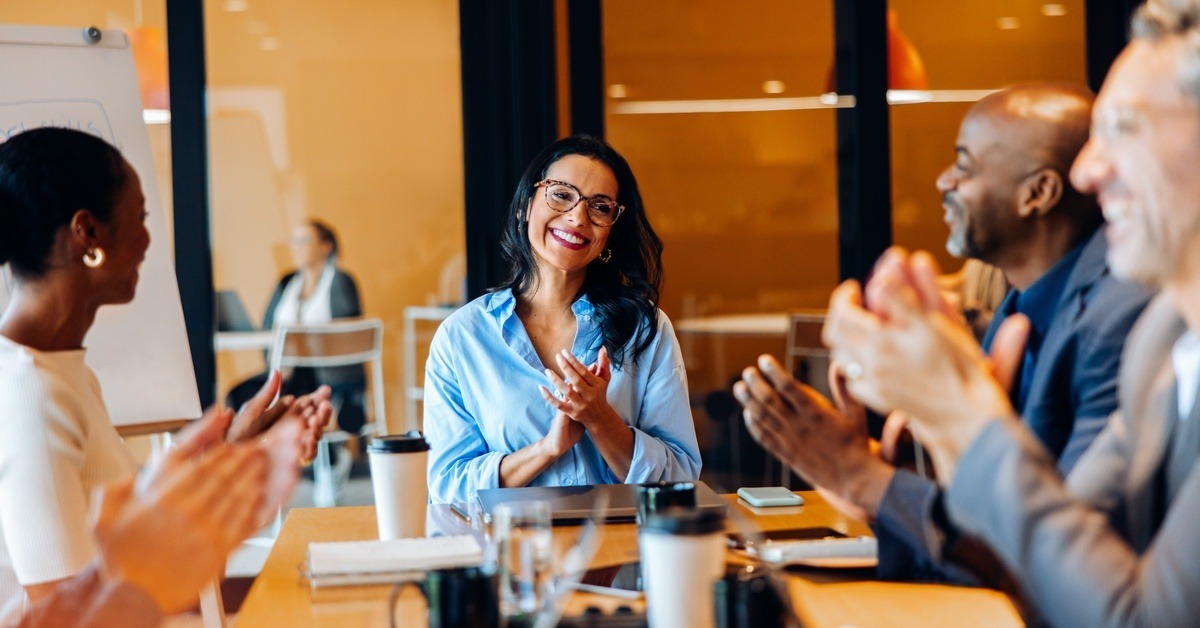 A smiling woman in a blue blouse sits and applauds alongside her coworkers at a meeting table in a glass-walled conference room.