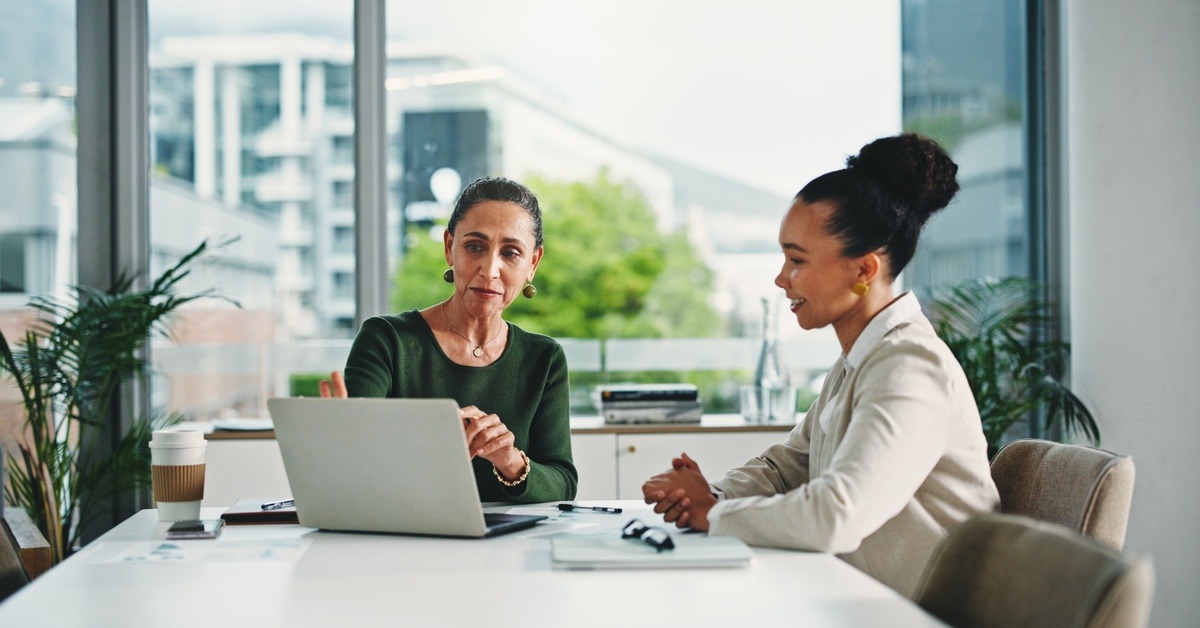 Two women review a laptop together while seated at a white table in a bright office with large windows and city views.