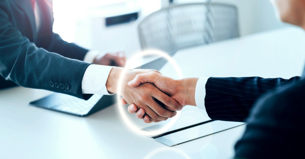 Two businesspeople shake hands across a desk in a bright office, their hands encircled with a glowing ring.