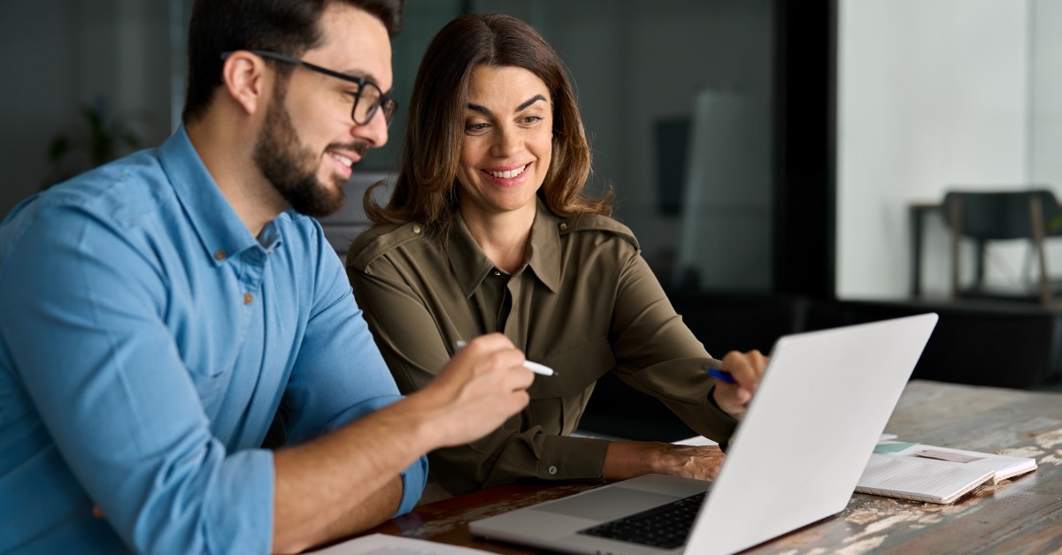 A smiling man and woman review a laptop together at a wooden table in an office with a glass wall behind them.