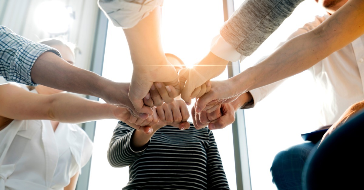 A group of coworkers join fists in a circle during a team huddle, illuminated by sunlight shining through the nearby window.
