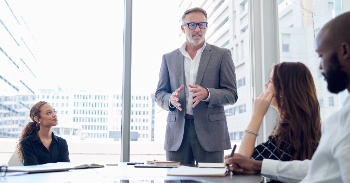 A man stands and speaks to colleagues around a conference table during a meeting in a bright office.