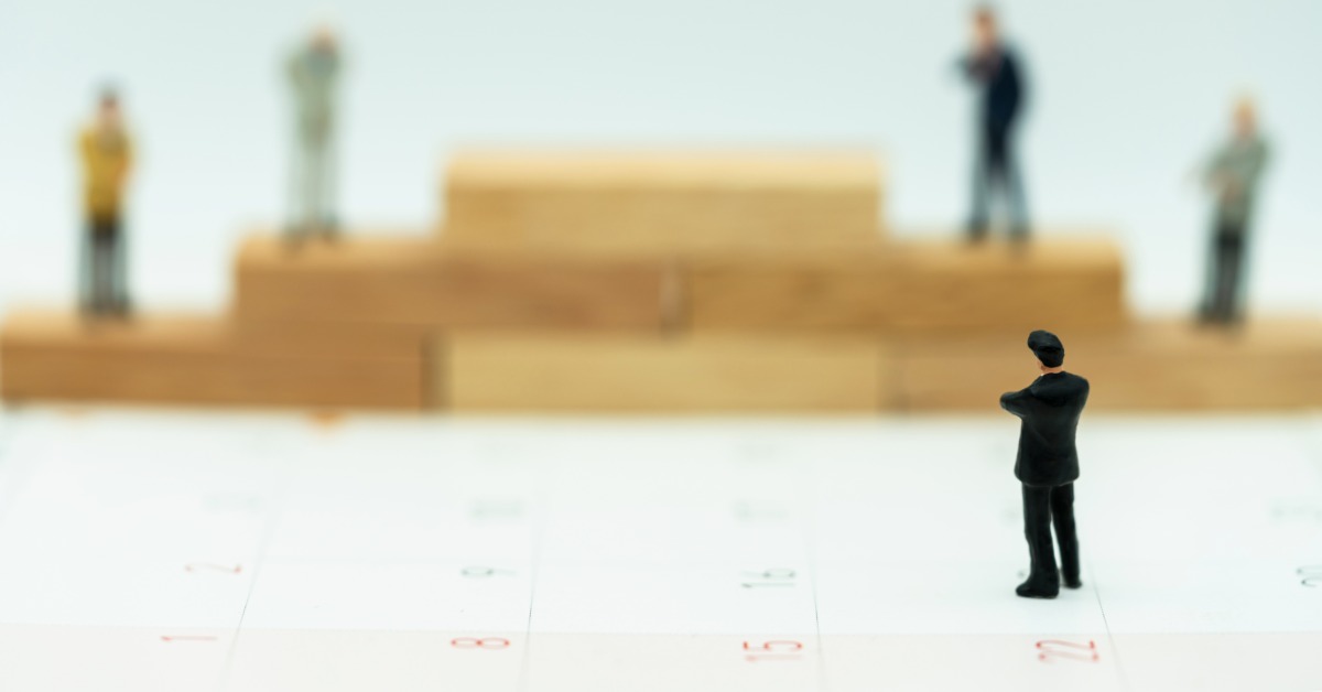 A mini business figure stands with his arms crossed, staring at a blurred podium with figures on every level but the top.
