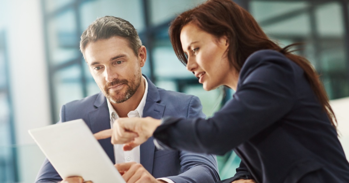 Two business professionals review a document together at a desk, with the woman pointing to a detail on the page.
