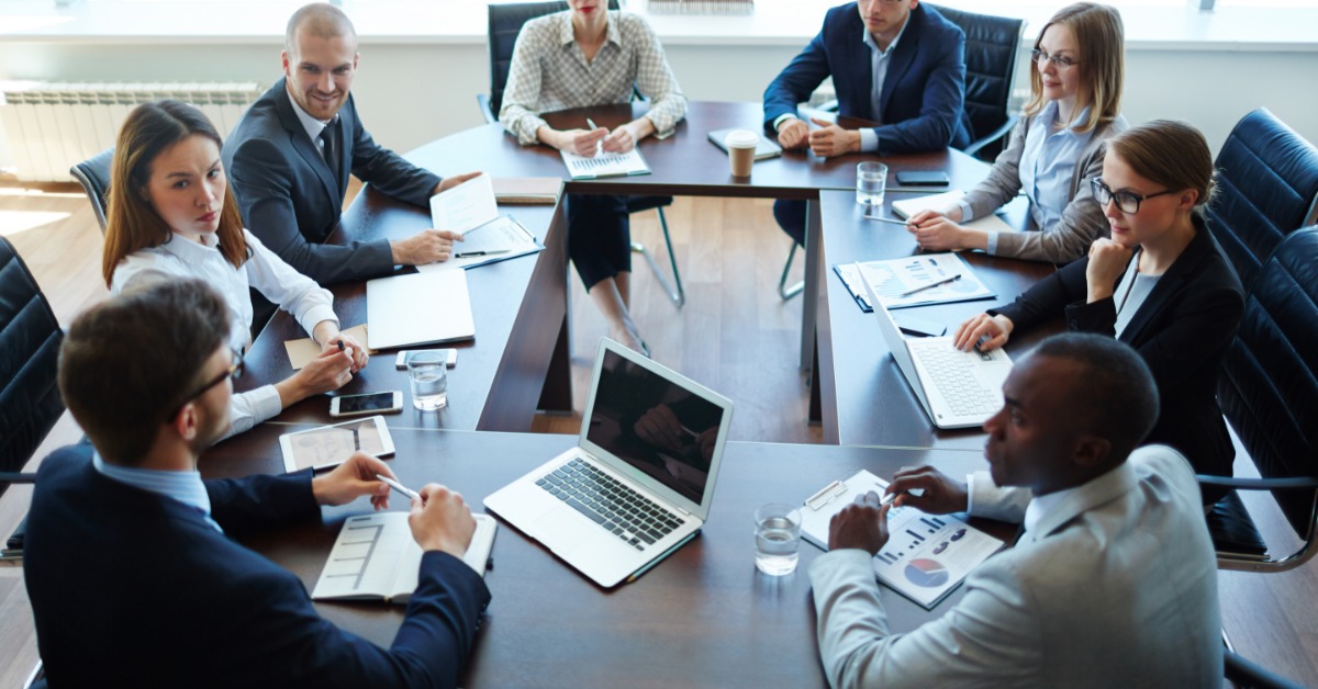 Eight professionally dressed executives sitting around a large wooden table in a company’s meeting room.