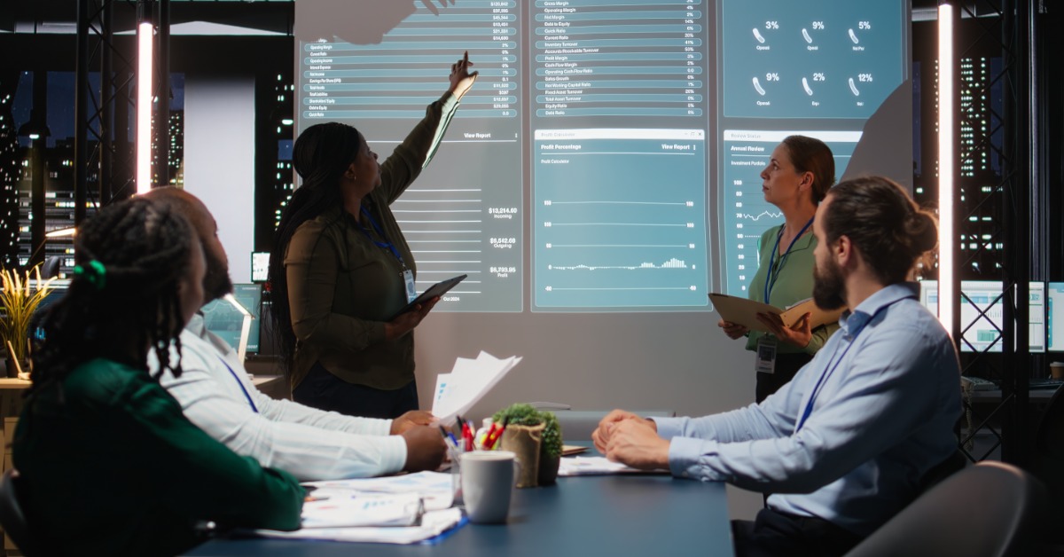 Three employees sitting at a table watching two other employees give a presentation with digital graphs.