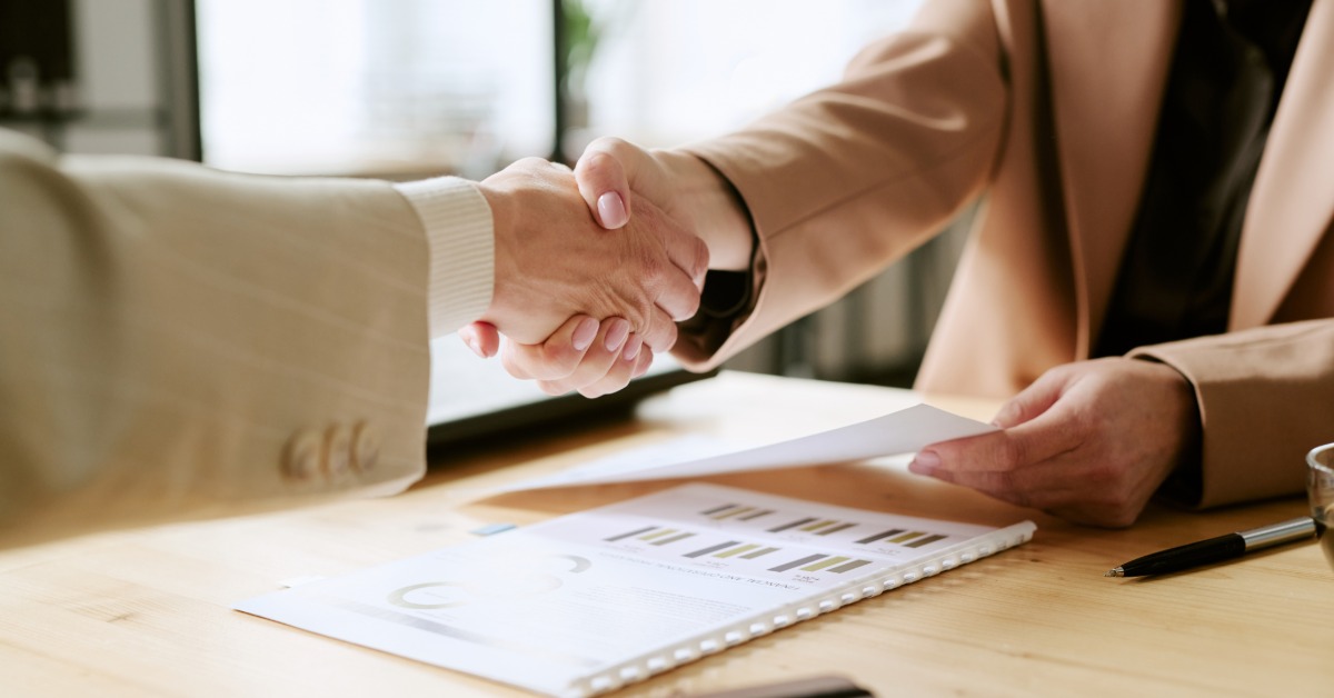 Two executives shaking hands over a round wooden table.