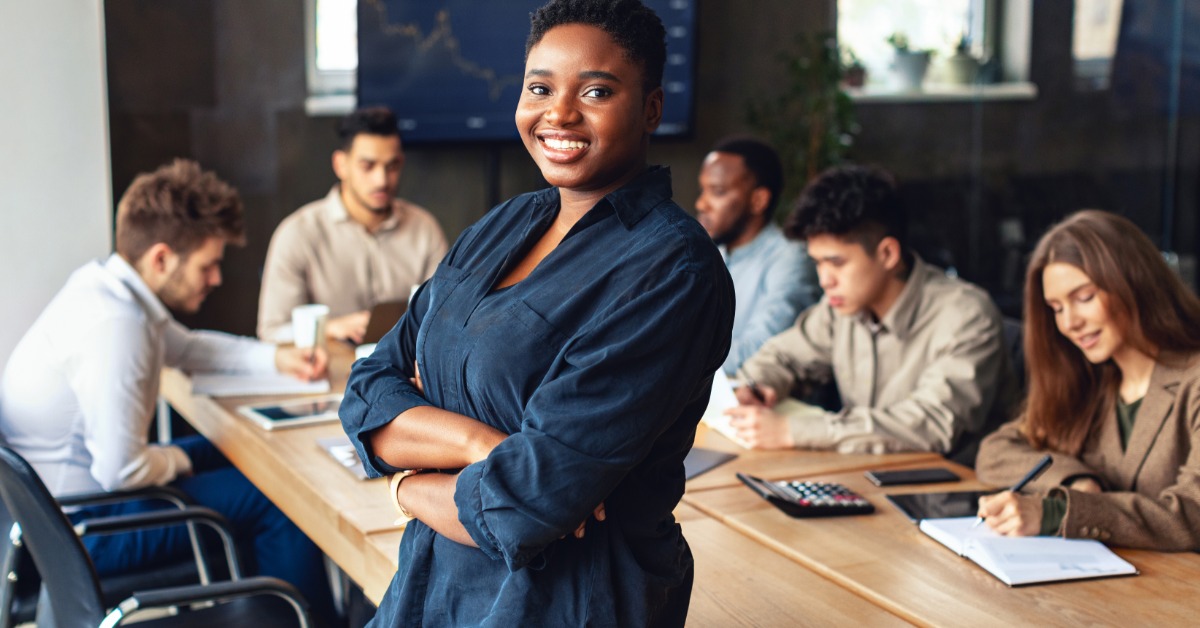 An executive learning against a table crossing their arms and smiling. Five employees sit at the table.