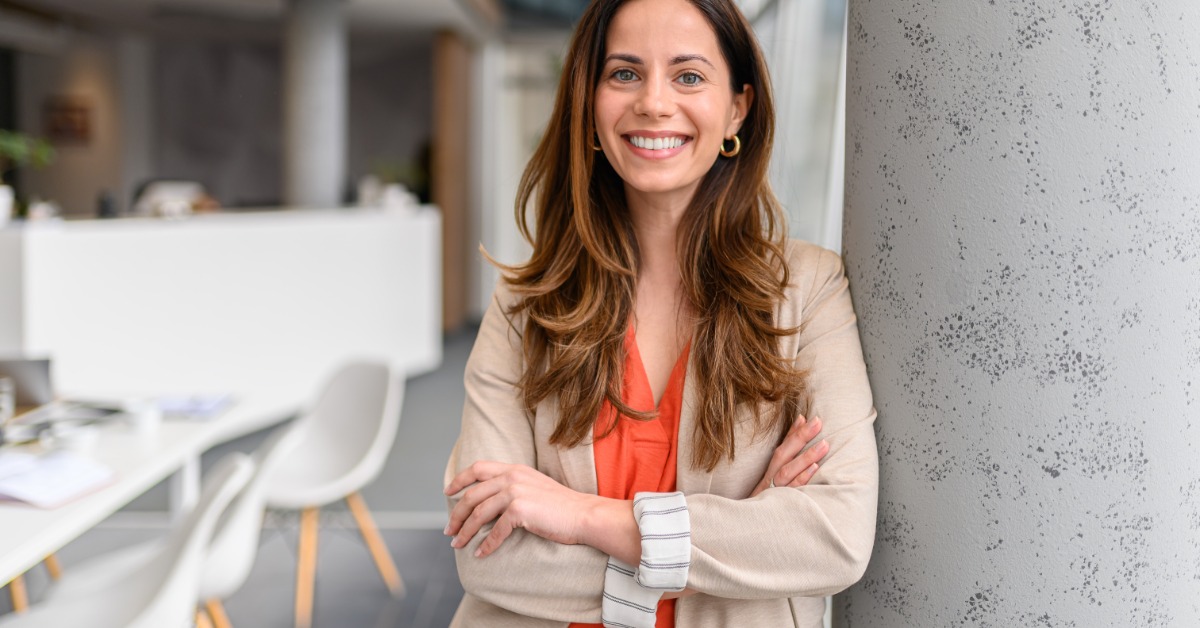 An executive crossing their arms and smiling. The executive leans against a tall pillar in their workplace.