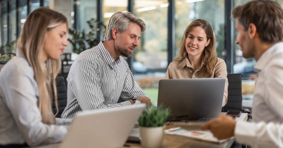 Four professionally dressed employees sitting at a table next to large windows. Two laptops sit on the table.