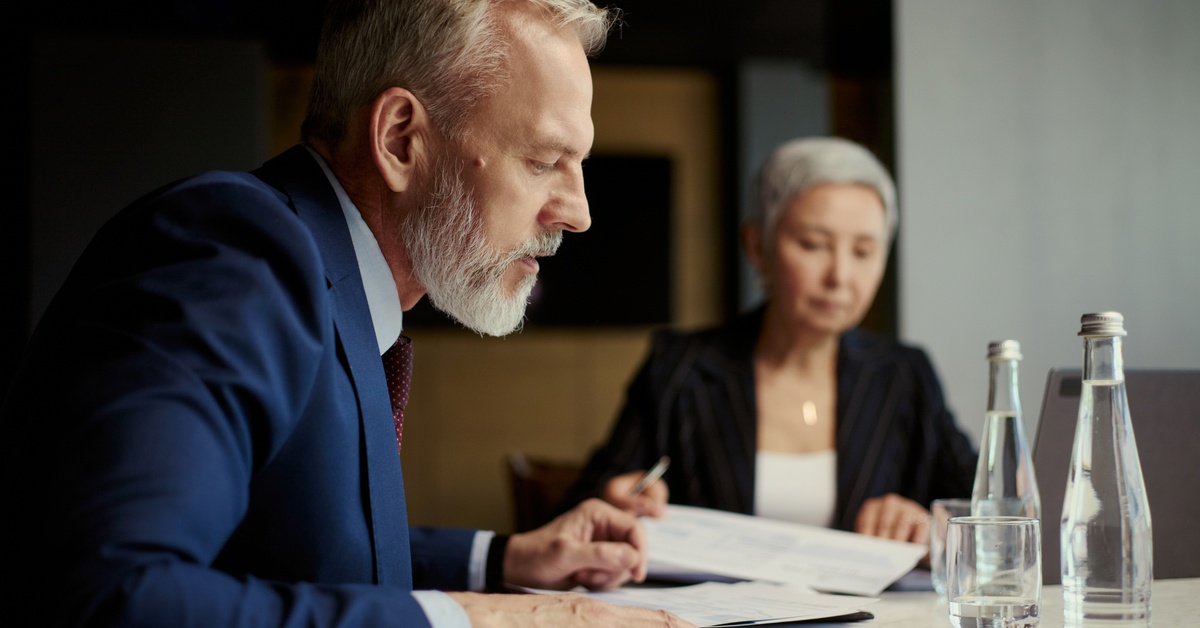 Two professionally dressed executives sit at a conference table. They both look at documents in front of them.