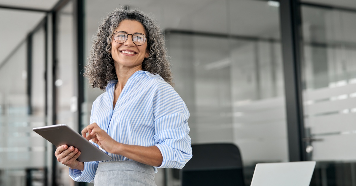 A professional smiles in an executive office. The professional wears glasses and holds a large tablet.