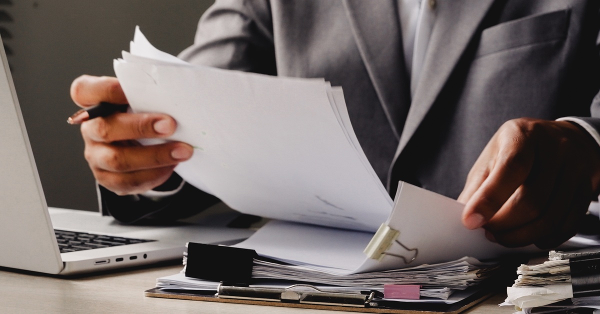 A person wearing a gray business suit sits at a desk and lifts several paper documents next to a laptop.