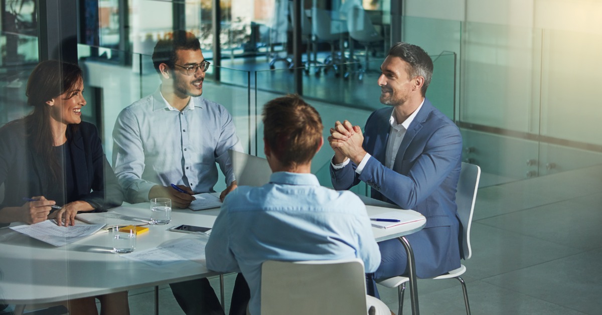 An executive sitting at the head of a round table talks to three employees sitting at the same table.