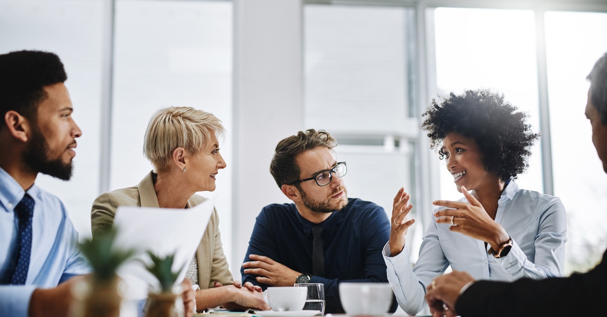 Five professionally dressed employees sitting around a table in an office. One employee talks and the other four listen.