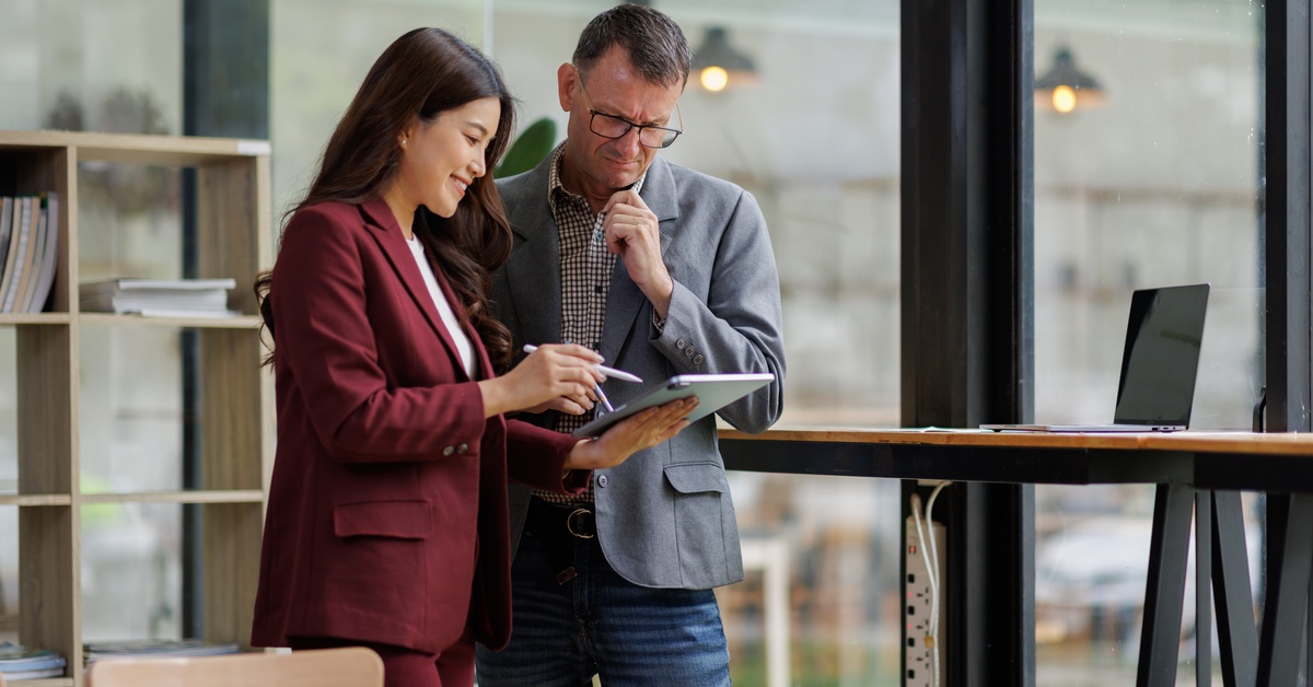 Two executives standing in a brightly lit office. They both look at a tablet that one of the executives holds.