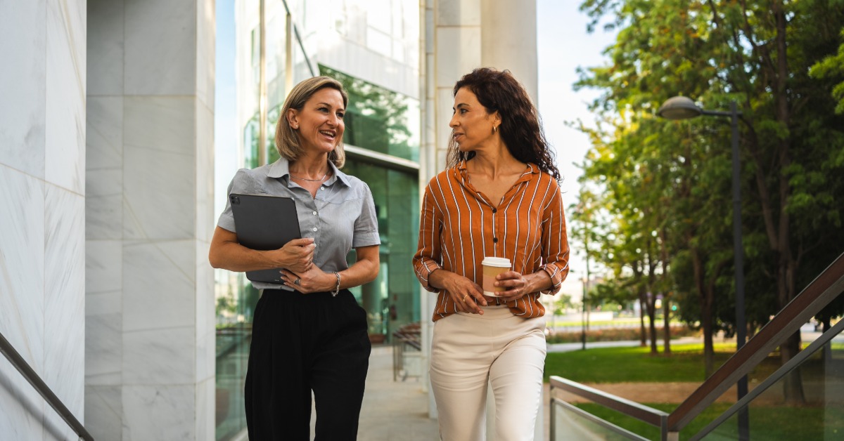 Two professionals walking outside during the daytime. One person holds a coffee cup and the other has a tablet.