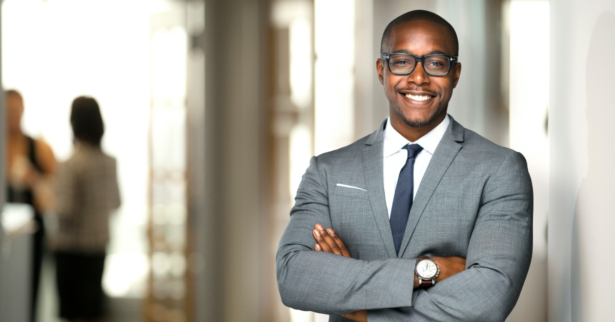 An executive wearing a gray suit and black glasses smiles in a hallway in an office. The executive crosses their arms.